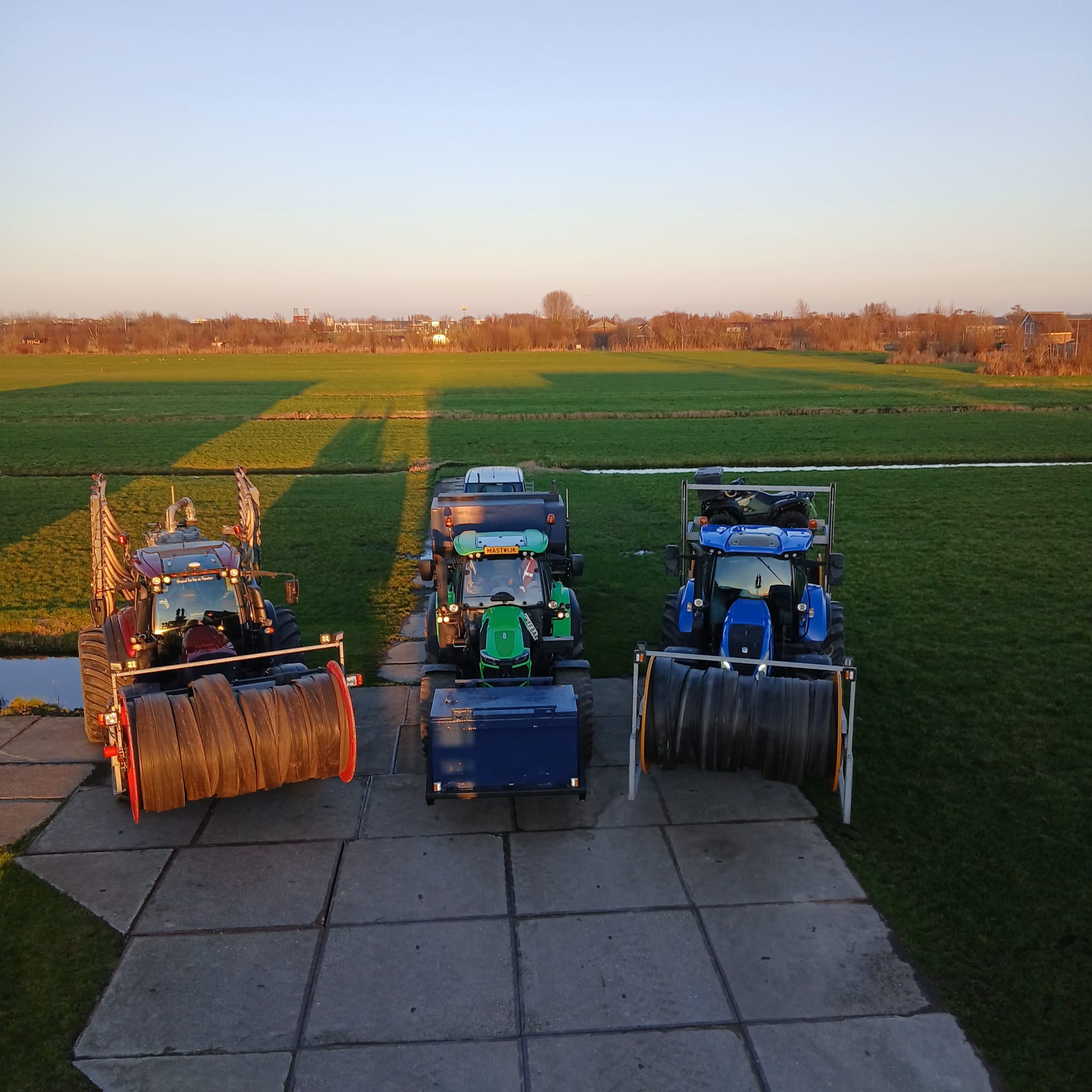 Machinepark van HarteWerkt met rode, groene en blauwe tractoren naast een weiland bij zonsondergang